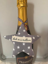 Load image into Gallery viewer, A softly padded star or heart with looped ribbon - so perfect for hanging around the bottle of something when gifting.  They can then choose where to hang it afterwards.  Choose your message - 'Dad in a Million', 'Grandad in a Million' or 'Happy Father's Day'  Fabric options are - grey stars, football, Union Jack or rugby - I have many more options so please use the 'Contact Us' page if you would like to discuss these.